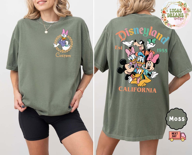 Puede incluir: Camiseta verde musgo con Daisy Duck en la parte delantera. La parte trasera de la camiseta muestra un colorido dise&ntilde;o de Disneyland con Mickey, Minnie, Donald y Goofy. Tambi&eacute;n se ve el texto "Disneyland California".