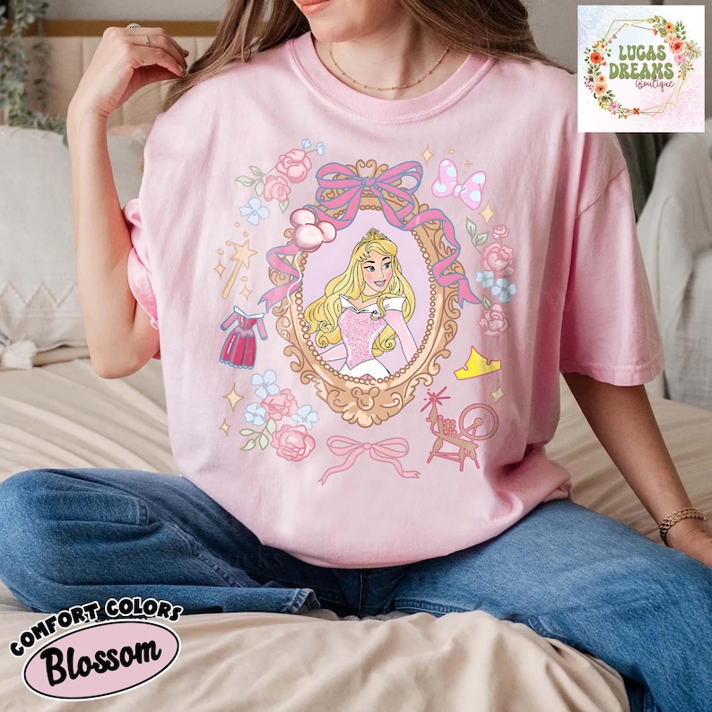 以下が含まれることがあります： ピンク色のComfort Colors Blossom Tシャツ。お姫様のイラストが額縁に入っています。ピンクのリボン、花、おとぎ話の要素がデザインされています。Tシャツは半袖です。