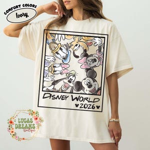 Op de afbeelding: Ivoor T-shirt met een ingelijste afbeelding van Disney-personages, waaronder Mickey Mouse, Donald Duck en Goofy. De tekst "DISNEY WORLD 2026" staat eronder. Het shirt heeft korte mouwen en een relaxte pasvorm.