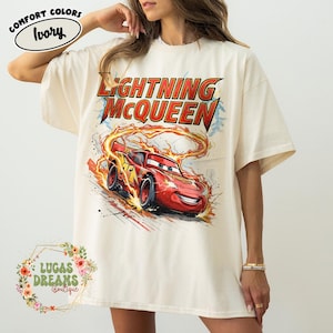 May include: Ivory-colored t-shirt featuring a graphic of Lightning McQueen from the Cars movie. The design includes the text "LIGHTNING McQUEEN" in red and yellow, with the race car in motion. The shirt also has the text "COMFORT COLORS" and "LUCAS DREAMS BOUTIQUE".