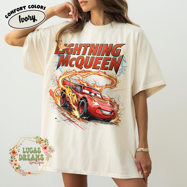 May include: Ivory-colored t-shirt featuring a graphic of Lightning McQueen from the Cars movie. The design includes the text "LIGHTNING McQUEEN" in red and yellow, with the race car in motion. The shirt also has the text "COMFORT COLORS" and "LUCAS DREAMS BOUTIQUE".