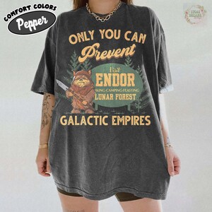 Puede incluir: Camiseta gris oscuro con el texto "ONLY YOU CAN Prevent Visit ENDOR LUNAR FOREST GALACTIC EMPIRES". La camiseta presenta un personaje de dibujos animados Ewok sosteniendo un cuchillo. La camiseta también tiene el texto "COMFORT COLORS Pepper".
