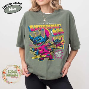 May include: Moss green t-shirt featuring a graphic of Stitch from Lilo & Stitch. The design includes the text "STITCH EXPERIMENT 626" in yellow and pink, with Stitch in a pink and blue outfit. The shirt also has the text "A COMPLETE MAYHEM!" and "HEAR THE CALL!"