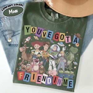 以下が含まれることがあります： トイ・ストーリーのキャラクターたちが描かれたモスグリーンのTシャツ。キャラクターのグループの上下には、「YOU'VE GOT A FRIEND IN ME」というテキストがカラフルなブロック体の文字でプリントされています。