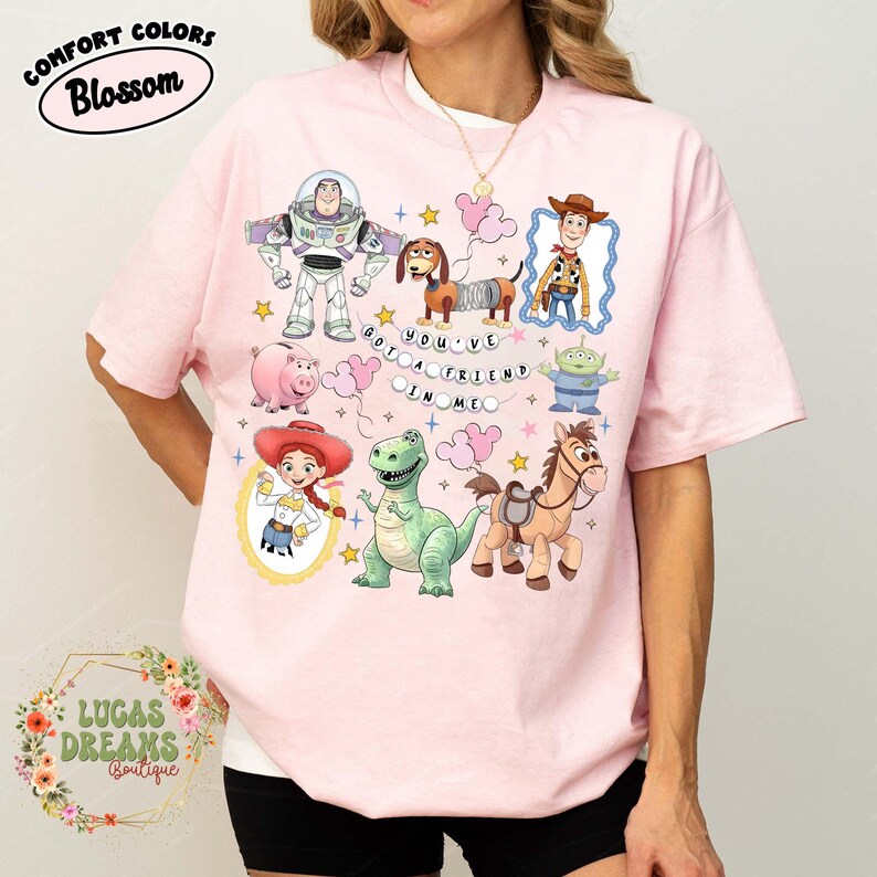 May include: Pink t-shirt featuring various Toy Story characters, including Buzz Lightyear, Woody, and Rex. The shirt has the phrase "YOU'VE GOT A FRIEND IN ME" and is adorned with stars and clouds. The shirt has the text "COMFORT COLORS Blossom" and "LUCAS DREAMS Boutique".