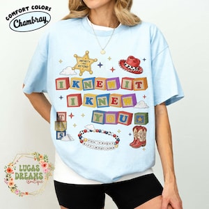 May include: A light blue t-shirt featuring a graphic print of colorful alphabet blocks spelling out "I KNEW IT I KNEW YOU". The design includes a sheriff star badge, a red cowboy hat, cowboy boots, and a beaded bracelet with the text "YOU'VE GOT A FRIEND IN ME".