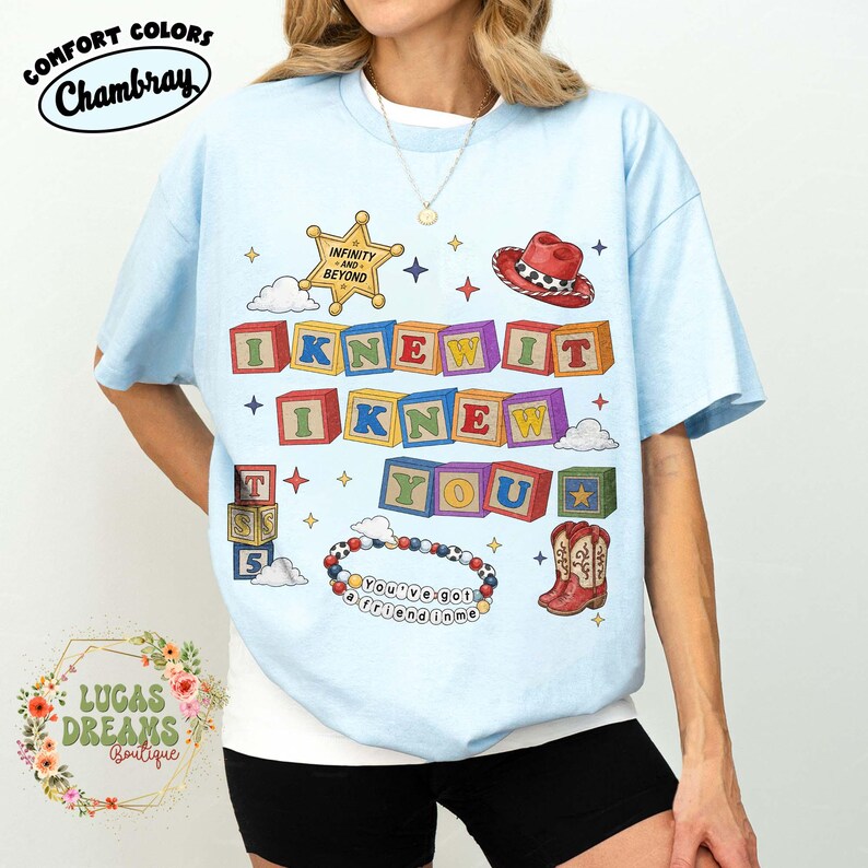 May include: A light blue t-shirt featuring a graphic print of colorful alphabet blocks spelling out "I KNEW IT I KNEW YOU". The design includes a sheriff star badge, a red cowboy hat, cowboy boots, and a beaded bracelet with the text "YOU'VE GOT A FRIEND IN ME".