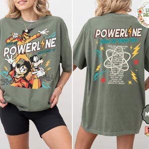 Puede incluir: Una camiseta verde musgo con un gr&aacute;fico de Powerline, Mickey y Goofy. La parte delantera de la camiseta muestra la palabra "POWERLINE" con personajes de dibujos animados. La parte trasera de la camiseta tiene "POWERLINE Stand Out World Tour" con una lista de ciudades.