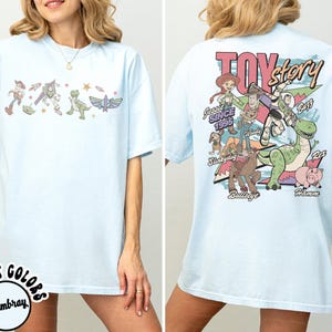 May include: Pale blue t-shirt with Toy Story characters on the front and back. The front shows Woody, Buzz Lightyear, and other characters. The back has a larger graphic with the words "Toy Story" and more characters.