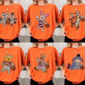 May include: Orange Halloween-themed t-shirts featuring various cartoon characters, including Chip and Dale, Piglet, Tigger, Winnie the Pooh, Roo, and Eeyore. Each character is dressed in a mummy costume and holding a jack-o'-lantern.