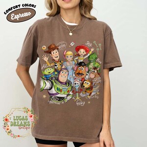 Op de afbeelding: Een espresso-bruin t-shirt met korte mouwen en een grafische print van diverse Toy Story-personages, waaronder Woody, Buzz Lightyear, Jessie, Rex, Bullseye, Mr. Potato Head, Forky en een alien, met handgeschreven namen van de personages.