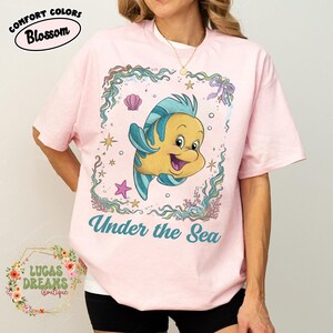 May include: Pink t-shirt with a cartoon illustration of a yellow and blue fish inside a decorative frame. The text "Under the Sea" is below the fish. The shirt also has the text "Comfort Colors Blossom" and "Lucas Dreams Boutique".