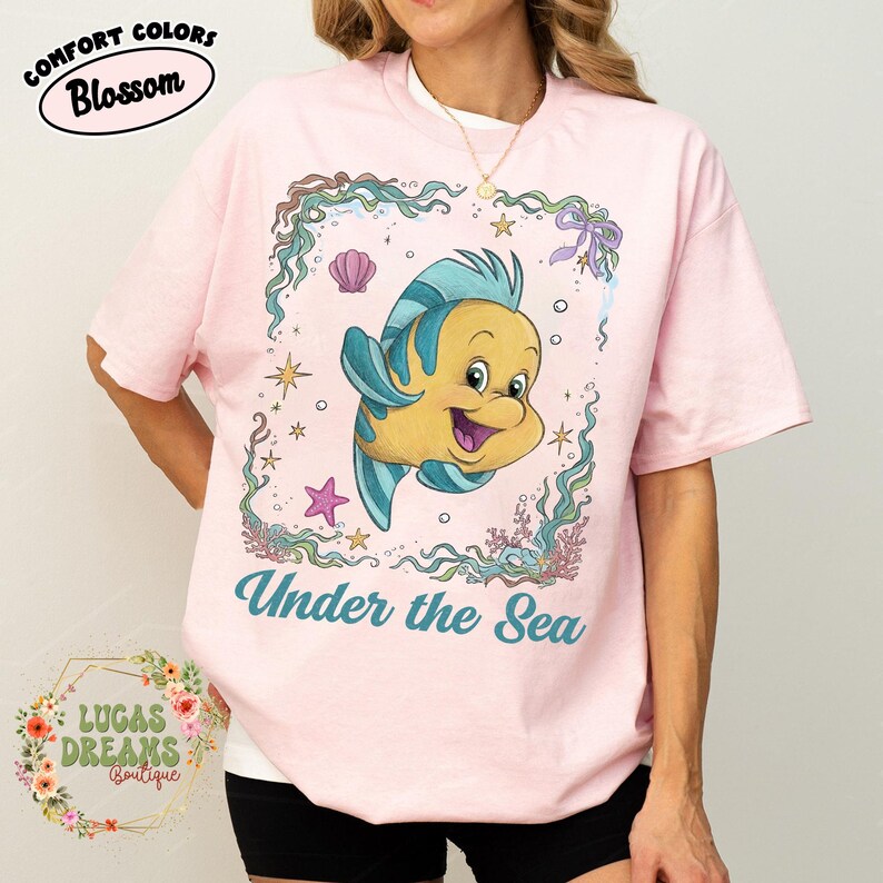 May include: Pink t-shirt with a cartoon illustration of a yellow and blue fish inside a decorative frame. The text "Under the Sea" is below the fish. The shirt also has the text "Comfort Colors Blossom" and "Lucas Dreams Boutique".