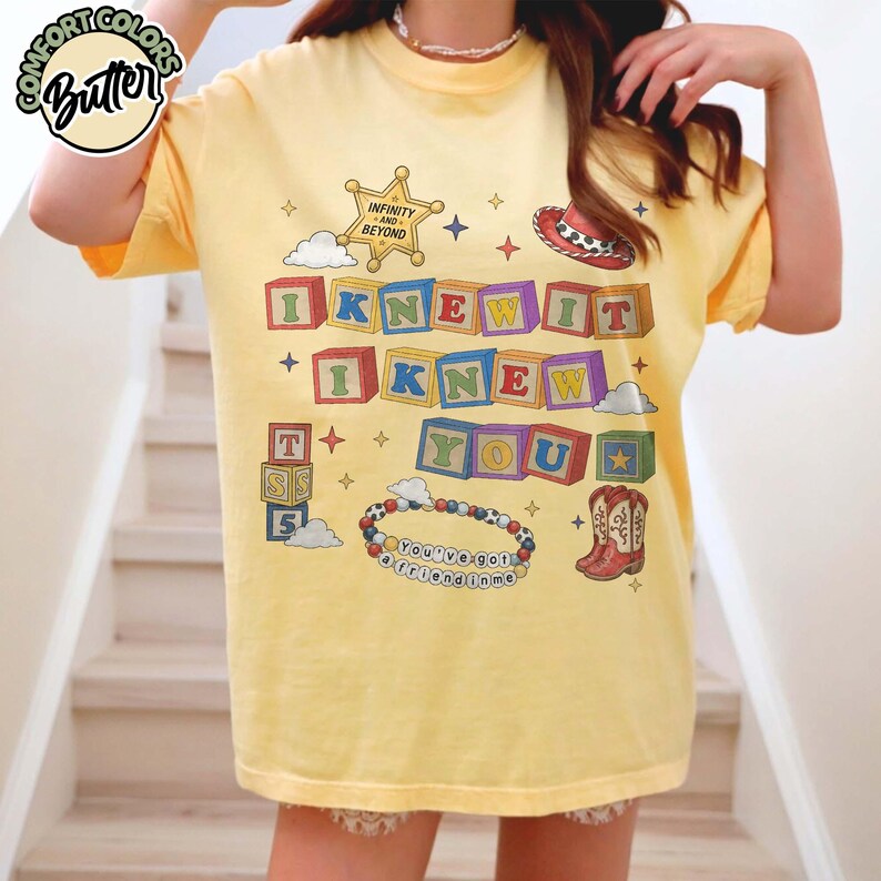 May include: A pale yellow t-shirt featuring a graphic print with colorful alphabet blocks spelling I KNEW IT I KNEW YOU. The design includes a sheriff badge with the text INFINITY AND BEYOND, a cowboy hat, boots, and a beaded bracelet with the text YOU'VE GOT A FRIEND IN ME.