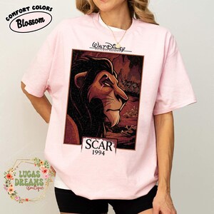 May include: A light pink t-shirt featuring a graphic print of the character Scar from The Lion King. The design includes the text Walt Disney Pictures Presents, Scar, and 1994. The illustration shows the lion in a dark, rocky landscape.