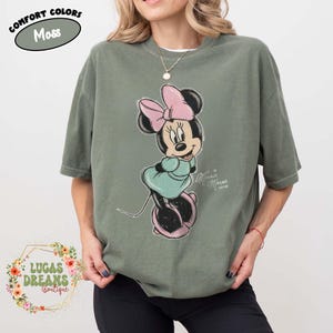 May include: A moss green short-sleeve t-shirt featuring a graphic of Minnie Mouse wearing a pink bow and a teal dress. The text "Minnie Mouse xoxo" is printed next to the character. A label in the top left corner reads "Comfort Colors Moss".