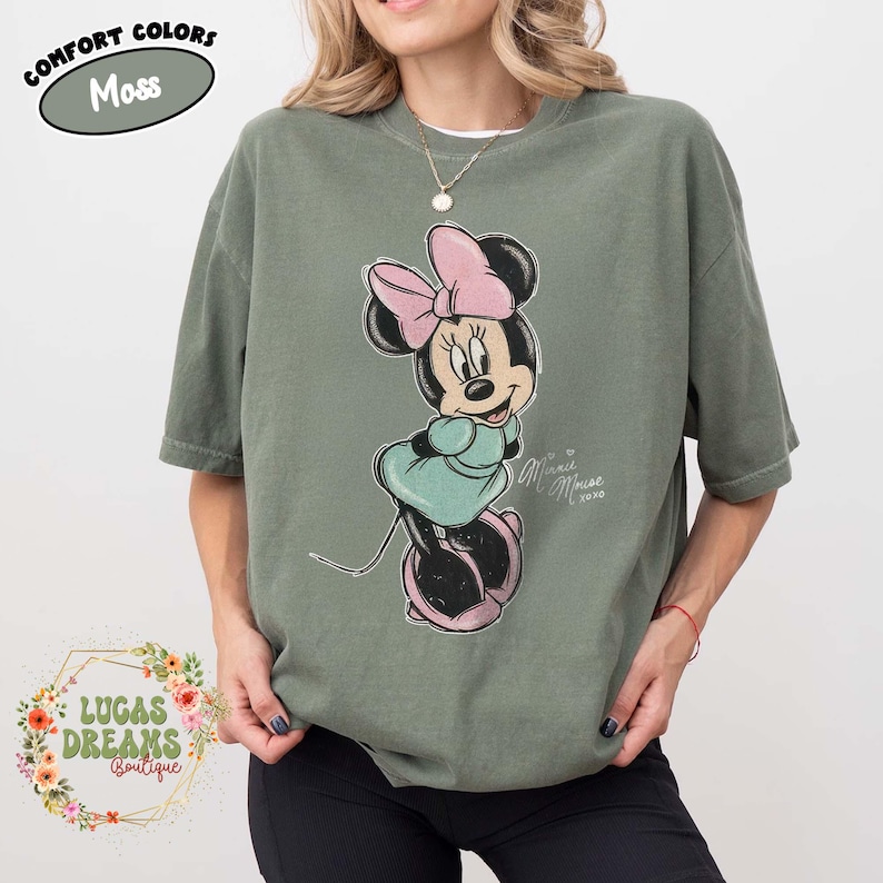 May include: A moss green short-sleeve t-shirt featuring a graphic of Minnie Mouse wearing a pink bow and a teal dress. The text "Minnie Mouse xoxo" is printed next to the character. A label in the top left corner reads "Comfort Colors Moss".