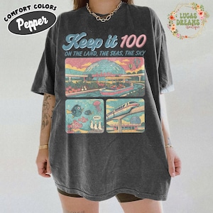 May include: Dark grey t-shirt with a colorful graphic featuring the text "Keep it 100 ON THE LAND, THE SEAS, THE SKY". The graphic includes illustrations of a boat, fish, a train, and a building. The shirt has the "COMFORT COLORS" and "Pepper" logos.