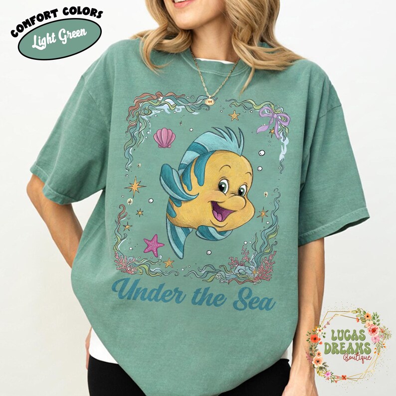 May include: Light green t-shirt featuring a cartoon fish with blue and yellow accents, surrounded by an underwater scene. The text "Under the Sea" is printed below the fish. The shirt also has the text "Comfort Colors" and "Light Green".