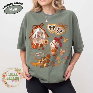 May include: Moss green t-shirt with a fall-themed graphic. The design includes a castle, pumpkins, Mickey and Minnie Mouse, Chip and Dale, and a pumpkin spice latte. The text "Comfort Colors" and "Lucas Dreams Boutique" are also visible.