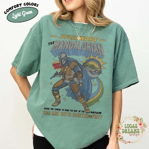 Puede incluir: Una camiseta de manga corta de color verde claro con un gr&aacute;fico de The Mandalorian y Grogu. El texto dice STAR WARS THE MANDALORIAN, BOUNTY HUNTER y WHEN YOU CHOOSE TO WALK THE WAY OF THE MANDALORE YOU ARE BOTH HUNTER & PREY. COMFORT COLORS est&aacute; impreso.
