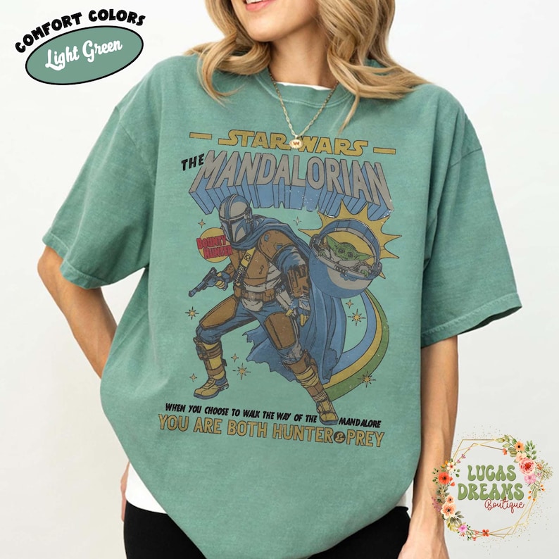 Puede incluir: Una camiseta de manga corta de color verde claro con un gr&aacute;fico de The Mandalorian y Grogu. El texto dice STAR WARS THE MANDALORIAN, BOUNTY HUNTER y WHEN YOU CHOOSE TO WALK THE WAY OF THE MANDALORE YOU ARE BOTH HUNTER & PREY. COMFORT COLORS est&aacute; impreso.