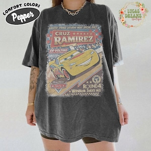 May include: Dark gray t-shirt featuring a graphic of Cruz Ramirez from the Disney Pixar Cars movie. The design includes the text "Radiator Springs Saturday Night Special Race" and "Round 4 Winner Takes All."