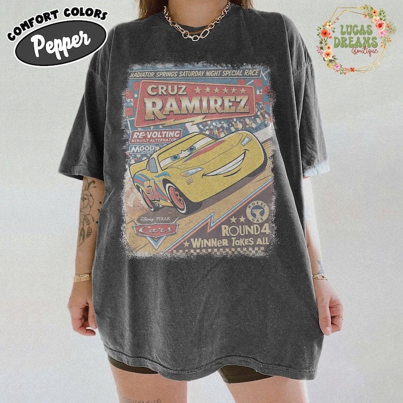 May include: Dark gray t-shirt featuring a graphic of Cruz Ramirez from the Disney Pixar Cars movie. The design includes the text "Radiator Springs Saturday Night Special Race" and "Round 4 Winner Takes All."