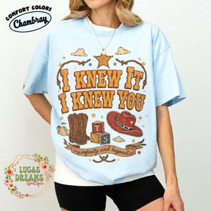 May include: A light blue t-shirt featuring a graphic print with the text I KNEW IT I KNEW YOU, a sheriff star, brown cowboy boots, alphabet blocks, a red cowboy hat, and a banner with the text infinity and beyond, all in a western style.