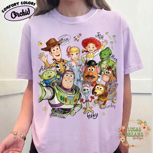 Op de afbeelding: Een lichtpaars t-shirt met korte mouwen en een grafische print van Toy Story-personages, waaronder Woody, Buzz Lightyear, Bo Peep, Jessie, Rex, Mr. Potato Head, Slinky Dog, Bullseye, Forky en een Alien, met hun namen in cursief schrift.
