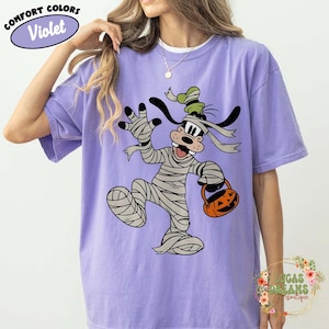 May include: Violet Comfort Colors t-shirt featuring a cartoon character dressed as a mummy holding a jack-o'-lantern bucket. The character has black ears and a green hat. The shirt has the words "Comfort Colors" and "Violet" in a circle.