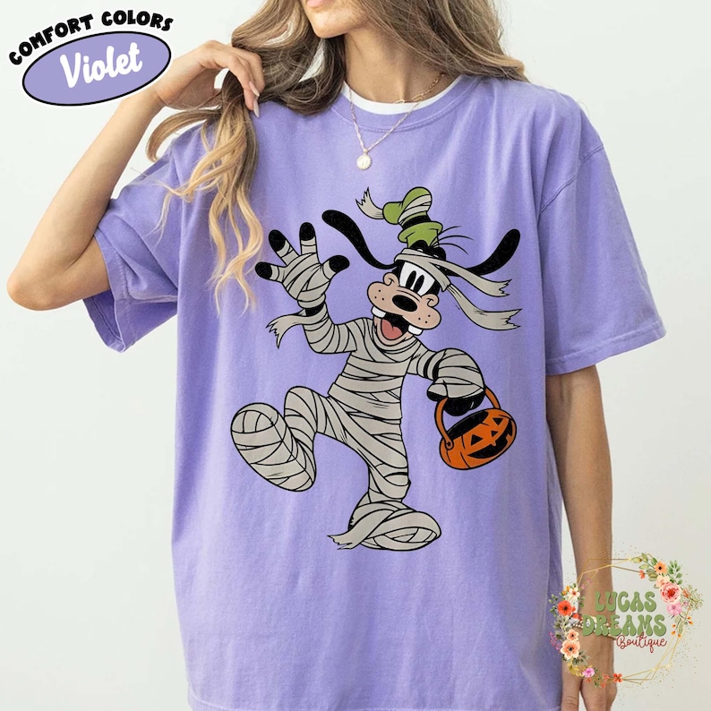 May include: Violet Comfort Colors t-shirt featuring a cartoon character dressed as a mummy holding a jack-o'-lantern bucket. The character has black ears and a green hat. The shirt has the words "Comfort Colors" and "Violet" in a circle.