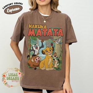 May include: Brown t-shirt featuring the phrase "Hakuna Matata" above a colorful graphic of characters from The Lion King. The shirt has a crew neck and short sleeves. The words "Comfort Colors Espresso" are printed on the top left.