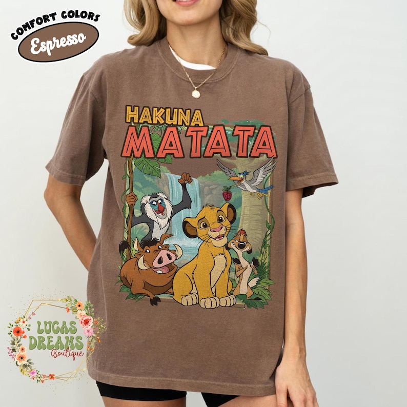 May include: Brown t-shirt featuring the phrase "Hakuna Matata" above a colorful graphic of characters from The Lion King. The shirt has a crew neck and short sleeves. The words "Comfort Colors Espresso" are printed on the top left.