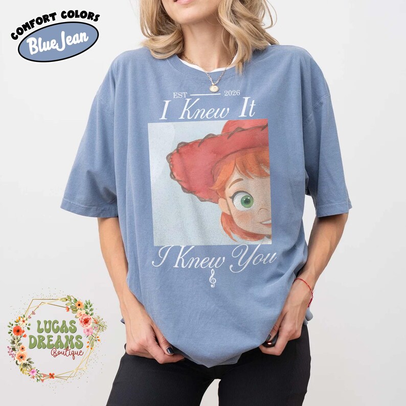 May include: A blue jean colored t-shirt featuring a graphic of a cartoon character with red hair and a red hat. The text reads "EST 2026 I Knew It I Knew You" with a small musical note symbol. The shirt has a crew neckline and short sleeves.