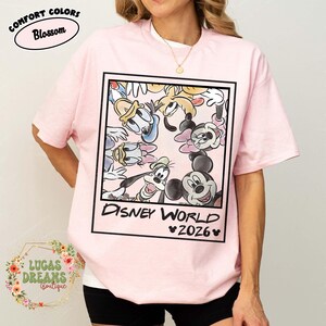 Op de afbeelding: Lichtroze T-shirt met een ingelijste afbeelding van Disney-personages, waaronder Mickey Mouse, Donald Duck en Goofy. De woorden "Disney World 2026" staan eronder. Het shirt heeft de woorden "Comfort Colors Blossom" linksboven.