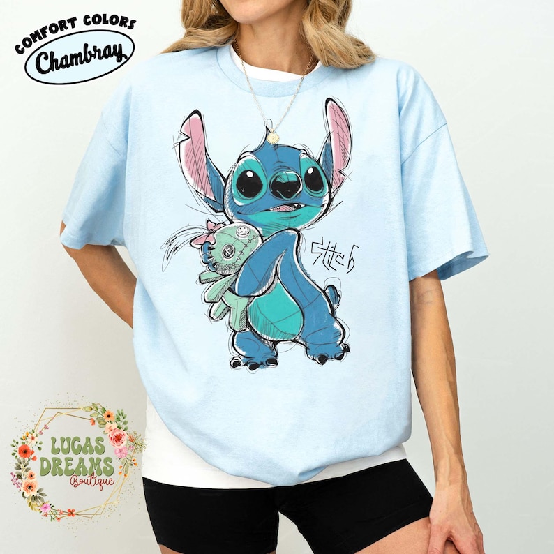 May include: A light blue short-sleeve t-shirt featuring a graphic illustration of the character Stitch holding a small doll. The design includes the text Stitch in a stylized script. The shirt has a crew neckline and a loose, relaxed fit.