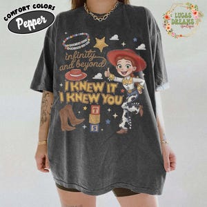 May include: A charcoal gray short-sleeve t-shirt featuring a graphic of a cowgirl character, a red hat, brown cowboy boots, and alphabet blocks. The text reads: You've got a friend in me, infinity and beyond, and I knew it I knew you.