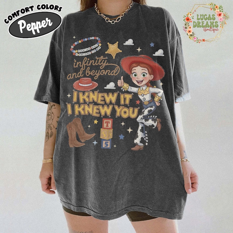 May include: A charcoal gray short-sleeve t-shirt featuring a graphic of a cowgirl character, a red hat, brown cowboy boots, and alphabet blocks. The text reads: You've got a friend in me, infinity and beyond, and I knew it I knew you.