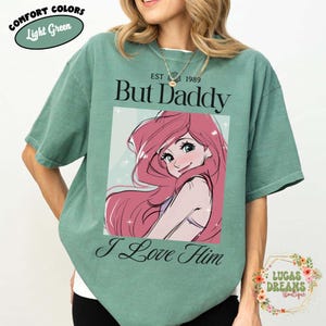 May include: Light green Comfort Colors t-shirt with the text "But Daddy I Love Him" and a cartoon illustration of a character with pink hair. The shirt also has the text "EST 1989" and "Lucas Dreams Boutique".