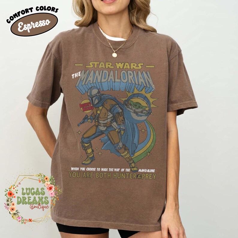 Puede incluir: Una camiseta de manga corta color marr&oacute;n espresso con un gr&aacute;fico de The Mandalorian y Grogu. El texto dice STAR WARS THE MANDALORIAN, BOUNTY HUNTER y WHEN YOU CHOOSE TO WALK THE WAY OF THE MANDALORE YOU ARE BOTH HUNTER & PREY.