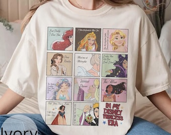 Pode incluir: Camiseta cor marfim com um design de grade apresentando ilustrações de princesas da Disney e texto. A camisa inclui imagens de Ariel, Aurora, Bela, Cinderela e outras. A frase "In My Disney Princess Era" também é exibida.