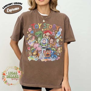 Pu&ograve; includere: T-shirt marrone con una grafica colorata dei personaggi di Toy Story, tra cui Woody, Buzz Lightyear e Rex. La parola "Toy Story" &egrave; scritta in un font giocoso. L'angolo in alto a sinistra riporta la scritta "Comfort Colors Espresso".