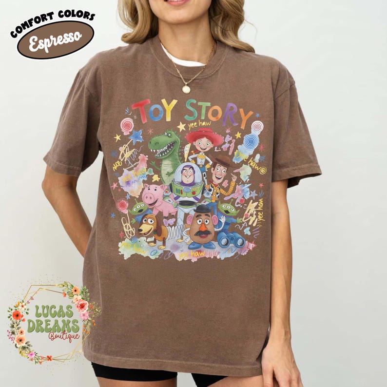 Pu&ograve; includere: T-shirt marrone con una grafica colorata dei personaggi di Toy Story, tra cui Woody, Buzz Lightyear e Rex. La parola "Toy Story" &egrave; scritta in un font giocoso. L'angolo in alto a sinistra riporta la scritta "Comfort Colors Espresso".