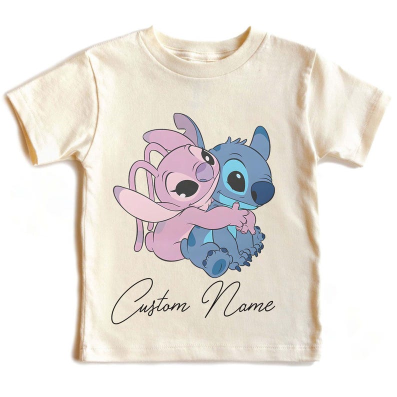 May include: Cream-colored t-shirt featuring a cartoon illustration of Stitch and Angel hugging. The characters are in shades of blue and pink. Below the image, the words "Custom Name" are written in a cursive font.