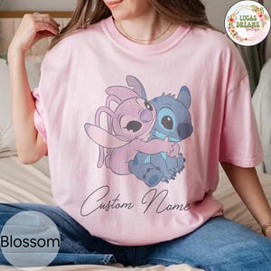 May include: A light pink t-shirt featuring a cartoon illustration of two characters hugging. One character is blue, the other is pink. The words "Custom Name" are written in cursive below the illustration. The word "Blossom" is in the bottom left corner.