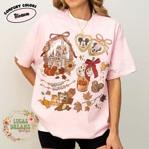 May include: A light pink t-shirt with a fall-themed graphic. The design includes a castle, pumpkins, Mickey and Minnie Mouse, Chip and Dale, and the words "Fall Vibes". The shirt has the text "Comfort Colors Blossom" and "Lucas Dreams Boutique".