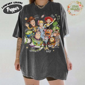 Op de afbeelding: Een antracietgrijs T-shirt met korte mouwen en een grafische print van Toy Story-personages, waaronder Woody, Buzz Lightyear, Jessie, Rex, Mr. Potato Head, Slinky Dog, Forky, Bo Peep, Bullseye en een Alien, met de namen in cursief schrift.