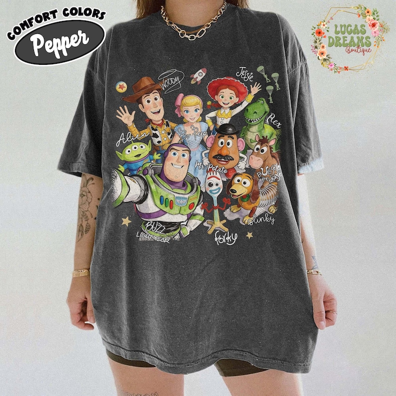 Op de afbeelding: Een antracietgrijs T-shirt met korte mouwen en een grafische print van Toy Story-personages, waaronder Woody, Buzz Lightyear, Jessie, Rex, Mr. Potato Head, Slinky Dog, Forky, Bo Peep, Bullseye en een Alien, met de namen in cursief schrift.