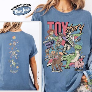 May include: Blue "Comfort Colors" t-shirt featuring a "Toy Story" graphic with characters like Woody, Buzz, and Jessie. The back of the shirt has a vertical design with more characters. The shirt is a light blue color.
